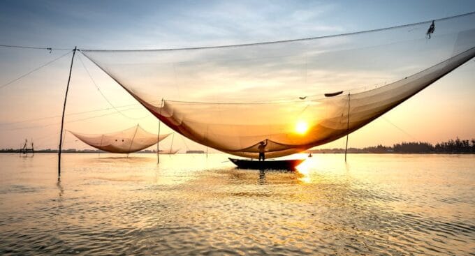 Fishing boat with large net is illuminated by the setting sun on a calm body of water. The sky is a blend of blue and orange hues.