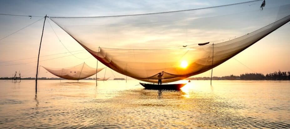 Fishing boat with large net is illuminated by the setting sun on a calm body of water. The sky is a blend of blue and orange hues.